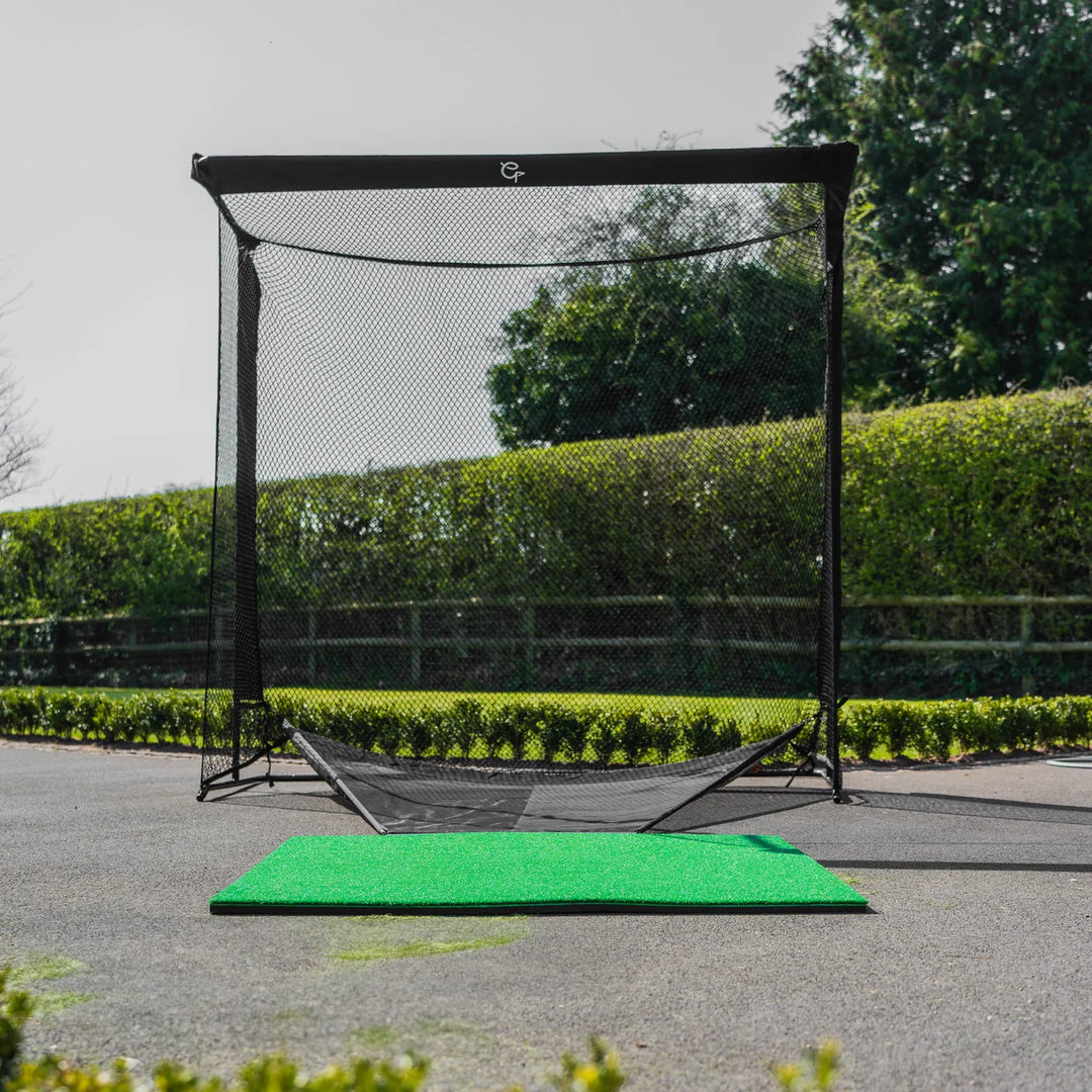 Golf practice net with a green mat on a paved area with trees in the background