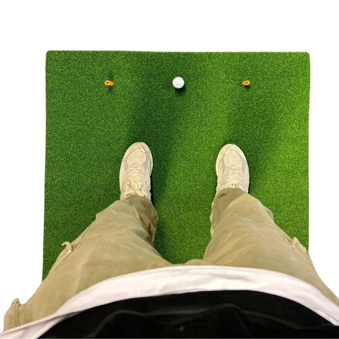 First-person view of a golfer standing on a green practice mat with a golf ball and two yellow tees placed ahead.