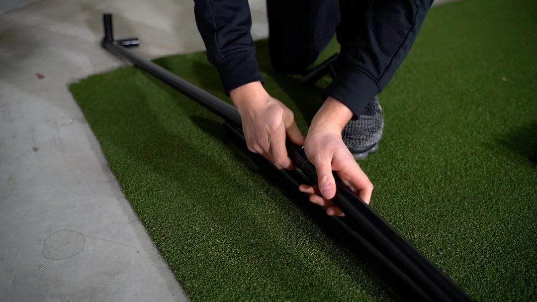 Hands assembling the frame of the GolfBays SimBox golf simulator enclosure on turf – available at Golf and Greens